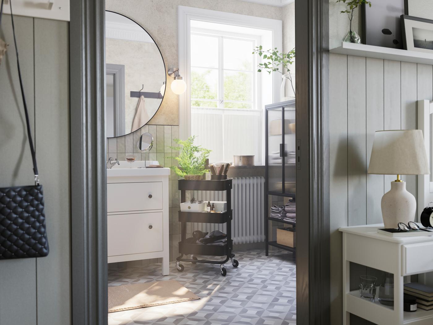 A grey bathroom with a big window has a white HEMNES washstand, a black RÅSKOG trolley and a MOSSJÖN glass door cabinet.