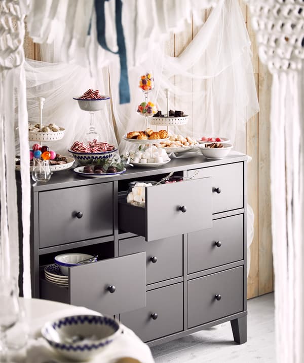 A grey chest of drawers carrying an array of stacked, filled and tiered plates and glasses overflowing with cookies and candy.