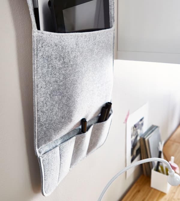 A grey fabric wall organiser hanging on a wall above a desk, with multiple pockets to hold pens and office supplies.