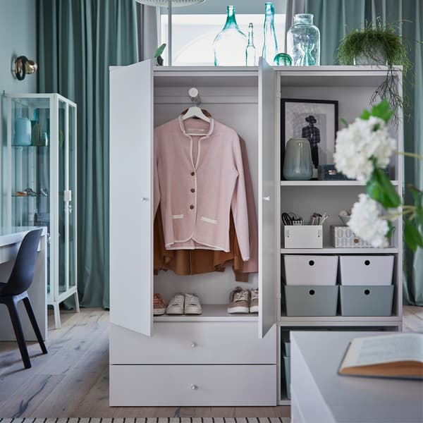 A grey/green bedroom with white clothes storage and storage shelves. The doors are open and show clothes hanging inside.