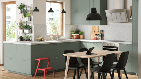 A grey-green kitchen, a wooden dining table, black chairs, a black pendant lamp and lots of herbs that hang on rails.
