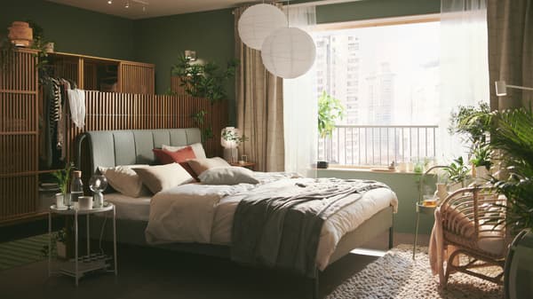 A grey-green TÄLLÅSEN upholstered bed frame with privacy screens behind it is in a serene green bedroom with large windows.