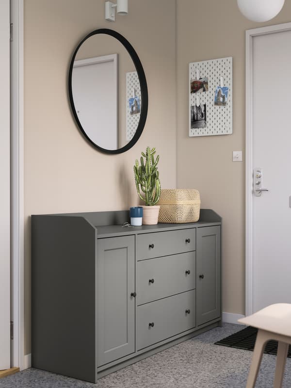 A grey HAUGA sideboard by a front door in a hallway, with a round mirror above it and a plant and a box on top.