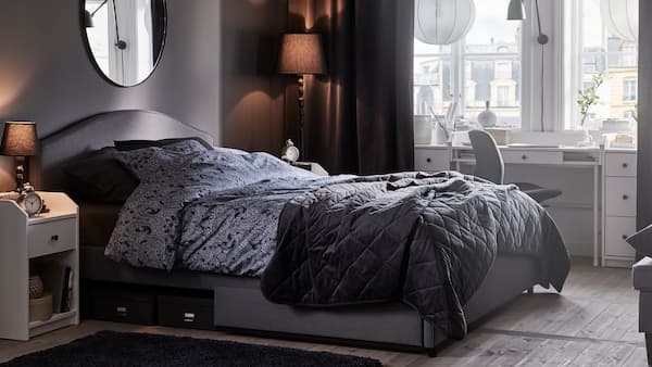 A grey HAUGA upholstered bed in a softly lit bedroom with a large window that has two white pendant lamps in front of it.