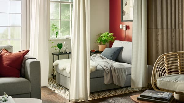A grey KIVIK chaise longue with a light grey INGABRITTA throw, behind white room-dividing GUNNLAUG sound-absorbing curtains.