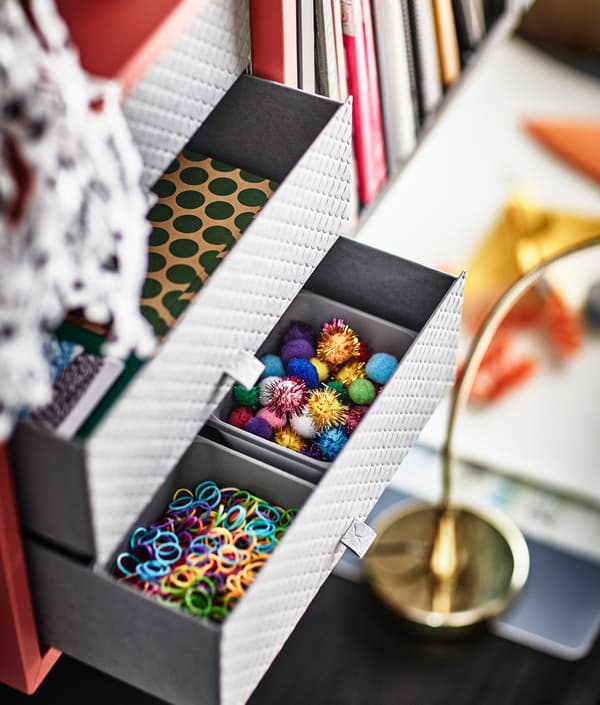 A grey paper desk organiser with 3 drawers, open to show organised and colourful rubber bands, sparkles and stationary.