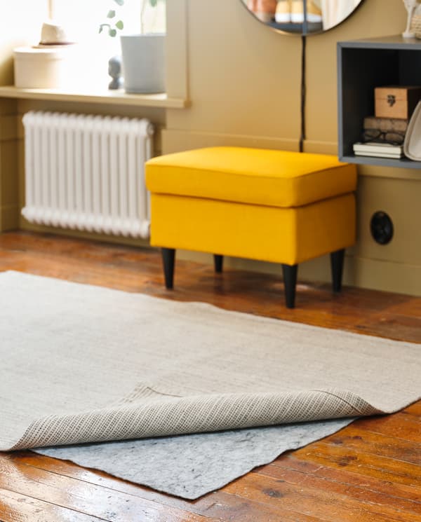 A grey rug underlay is under a natural/off-white rug. A yellow footstool and dark grey wall cabinet are in the background.