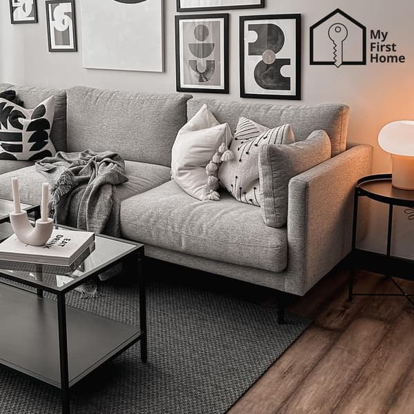 A grey SÖDERHAMN sofa is placed on top of grey rug in the living room, in front of a rectangular VITTJSÖ coffee table. Pictures in black frames are hung on the wall behind the sofa. Beside the sofa, a DEJSA table lamp placed on a black GLADOM table is turned on. The top right corner shows the "My First Home" icon, which consists of two parts: an outlined key placed on the outlined silhouette of a house, and the text "My First Home" placed on the right of the graphics.