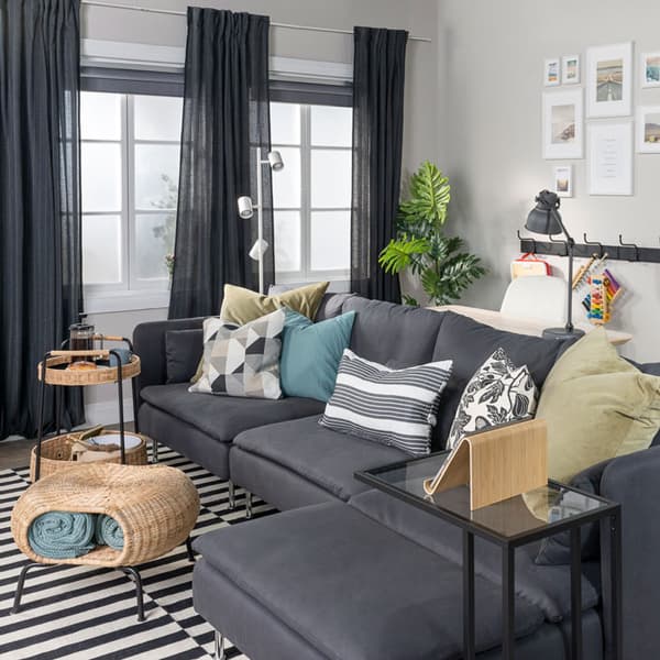 A grey sofa with a chaise is placed in the middle of a living room with open curtains. Beside the sofa is a laptop stand, footstool with storage, and a cart holding food.