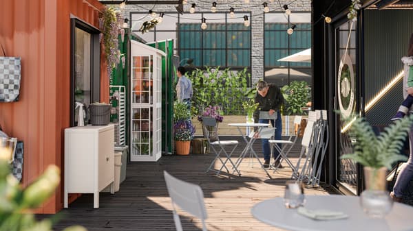 A grey SUNDSÖ chair is being set in an outdoor café area, adorned with plenty of plants, by a person wearing a black shirt.