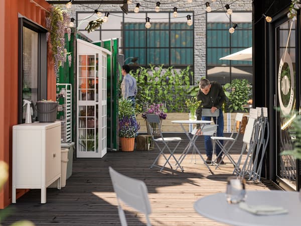 A grey SUNDSÖ chair is being set in an outdoor café area, adorned with plenty of plants, by a person wearing a black shirt.