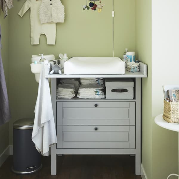 A grey SUNDWIK changing table with lots of baby accessories like pampers stacked on top and within the shelves.