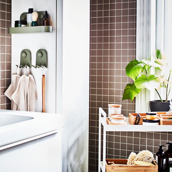 A grey-tiled bathroom with a white wash-stand, a white high cabinet, a white trolley with trays and jars plus a potted plant.