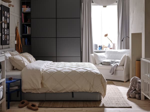 A grey upholstered bed covered in NYPONLUGGMAL bed linen in front of a dark grey PAX/MEHAMN wardrobe and a white armchair.