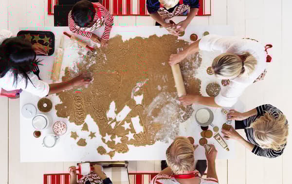 A group of children and adults amid gingerbread baking centred around a very large sheet of dough on a kitchen table.