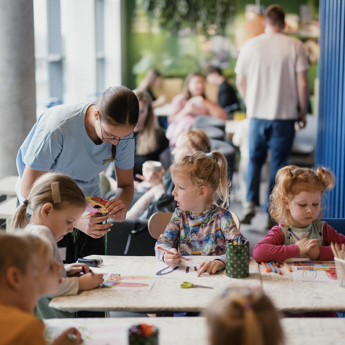 A group of children sit around a table at the Swedish Restaurant, drawing with coloured pencils while an adult leans over to help them.