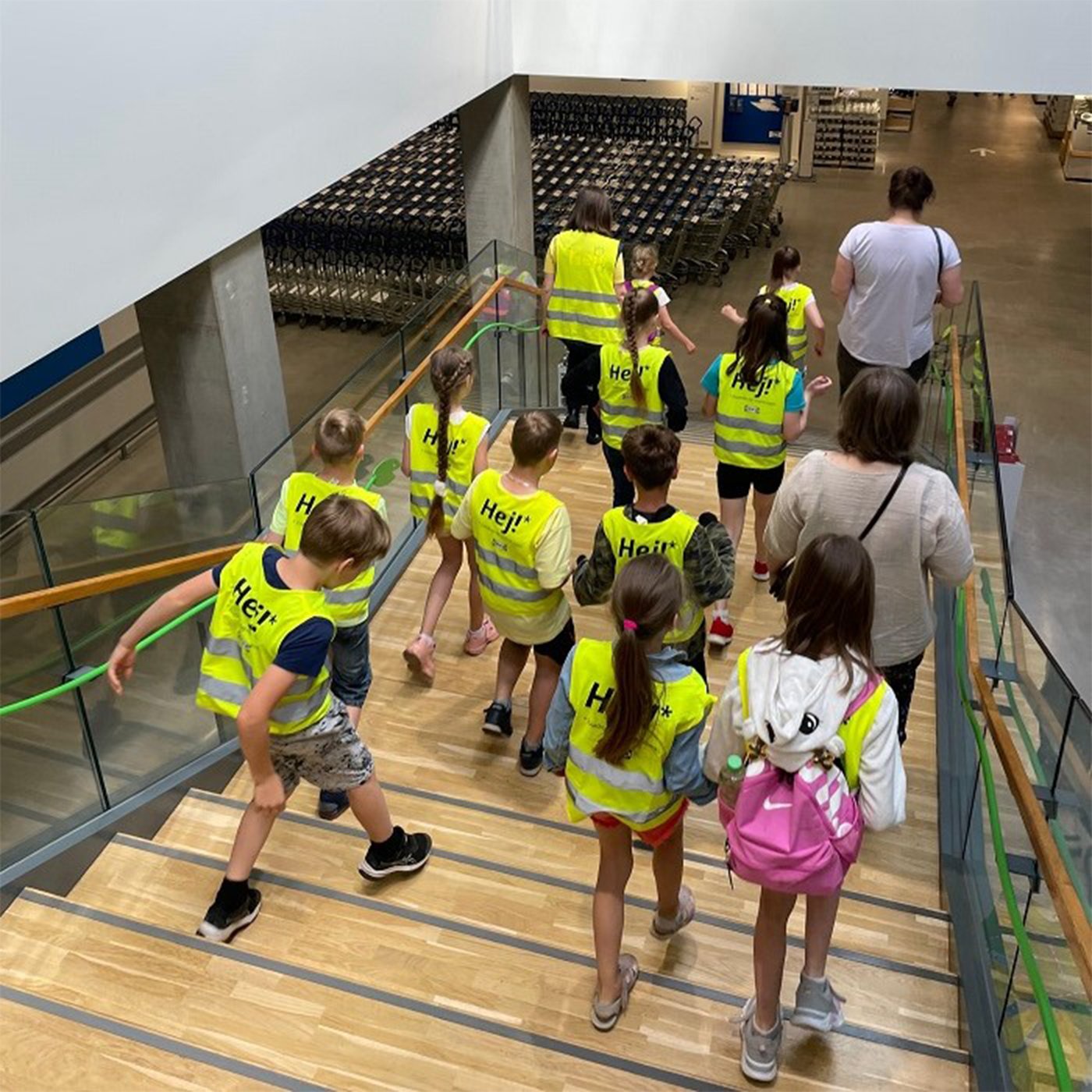A group of children wearing reflective vests, accompanied by two adults, climb down a wide wooden staircase.