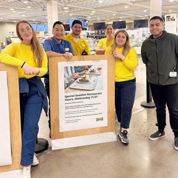A group of IKEA co-workers standing around an event sign for their store and smiling at the camera.