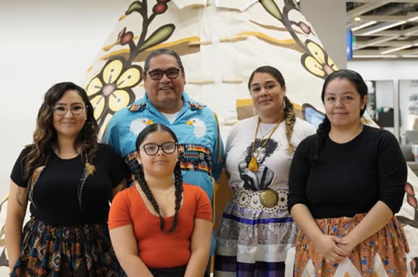 A group of people are gathered together smiling for a picture standing in front of a tipi. 