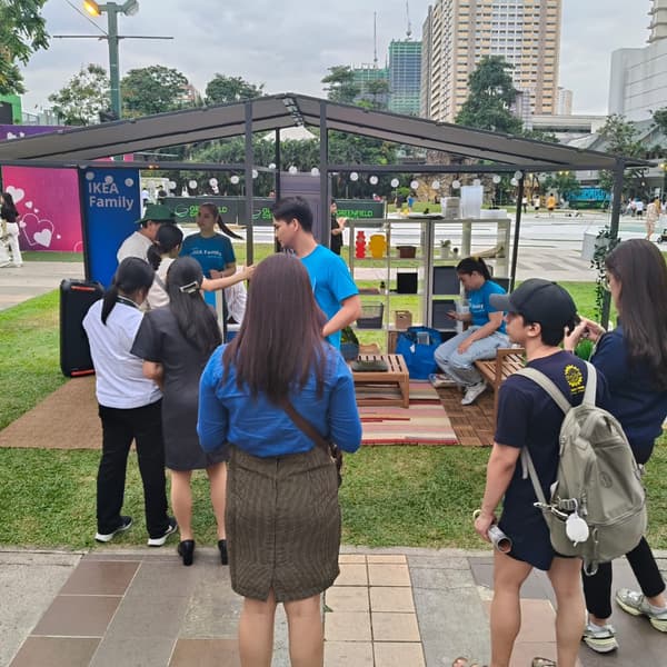 A group of people interacts around an IKEA booth showcasing furniture, with a park background and city buildings visible.