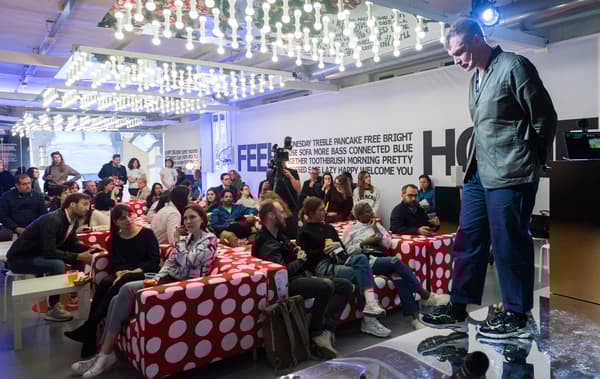 A group of people sit on red and white sofas in a large room with ceiling lights, listening to a man standing on a stage.