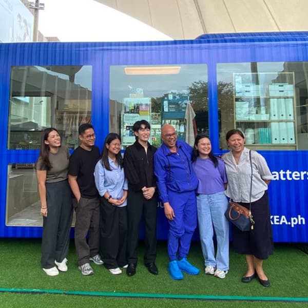 A group of seven people is smiling and standing close together, posing in front of a blue storefront with large windows. The scene conveys a cheerful and friendly atmosphere.