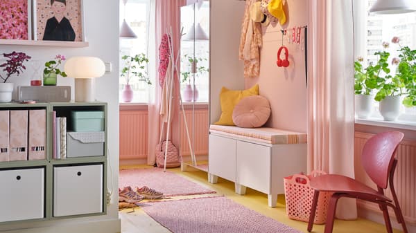 A hallway with a PLATSA mirrored wardrobe, a bench with pink and yellow cushions, two pink rugs and hanging hats and scarves.