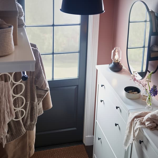 A hallway with two white shoe cabinets with various items on top, plus a white hat rack and a black mirror with storage.