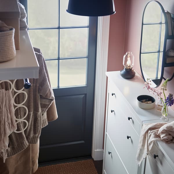 A hallway with two white shoe cabinets with various items on top, plus a white hat rack and a black mirror with storage.