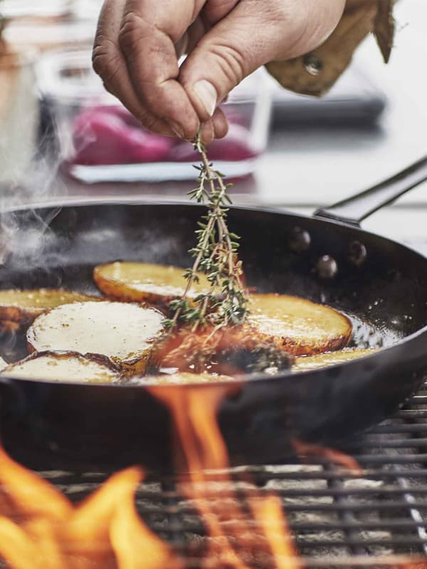 A hand adding a piece of rosemary into a VARDAGEN frying pan in steel on the grill/stove