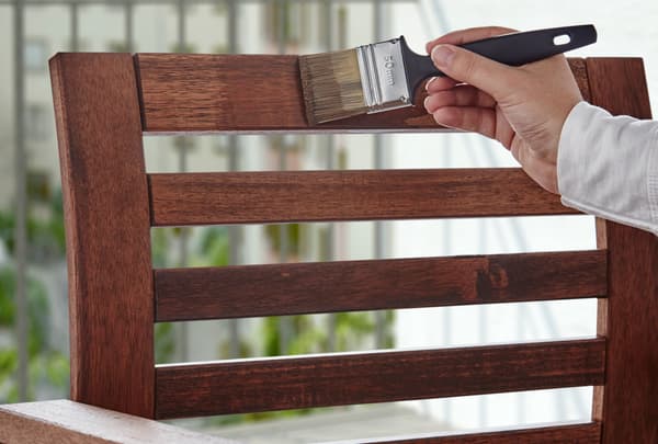 A hand holding a brush, covering the stained backrest of a wooden chair outdoors.