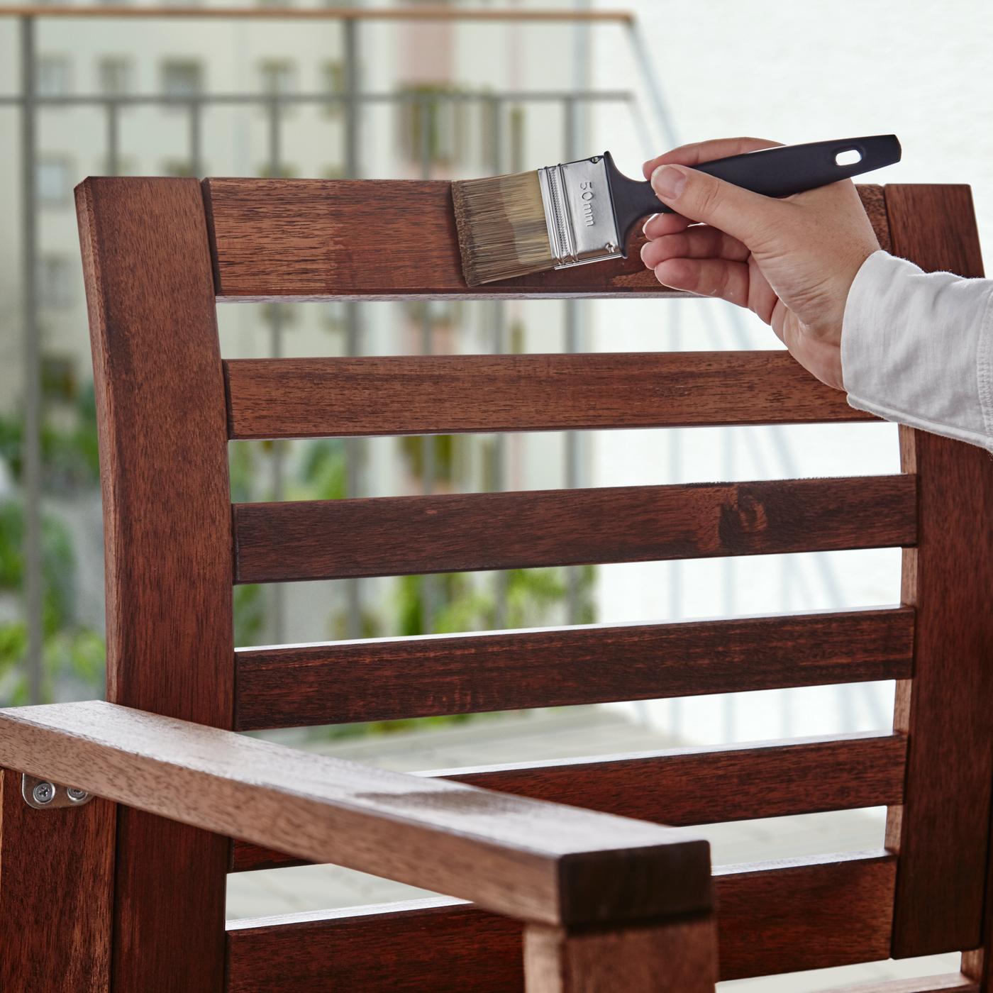 A hand holding a paint brush, covering the back of a wooden outdoor chair with stain.