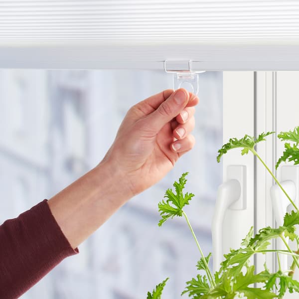 A hand holding the transparent plastic handle of a white cordless blind in a window with a green plant.