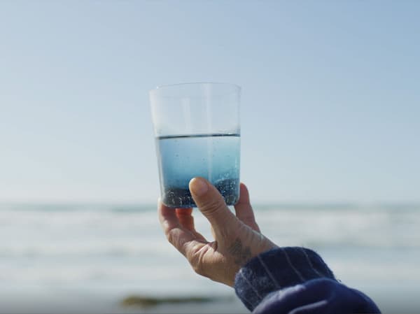 A hand holding up a glass of water in front of a sea in the background.