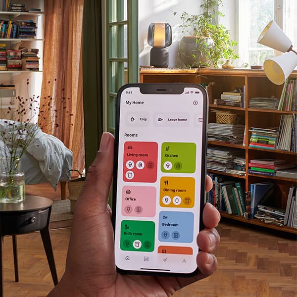 A hand holding up the IKEA Home smart app in front of a living room showing a screen with different options.