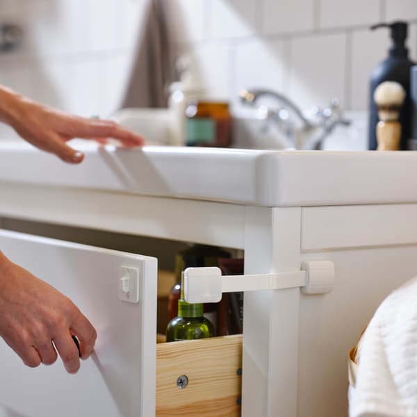 A hand holds open the drawer of a white HEMNES sink cabinet, attached to which is a white UNDVIKA multi-latch.