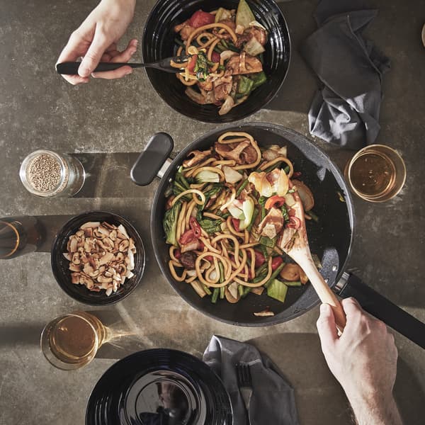 A hand is using a wooden spoon to serve a vegetable and noodle dish out of a MIDDAGMAT wok into two black bowls next to it.