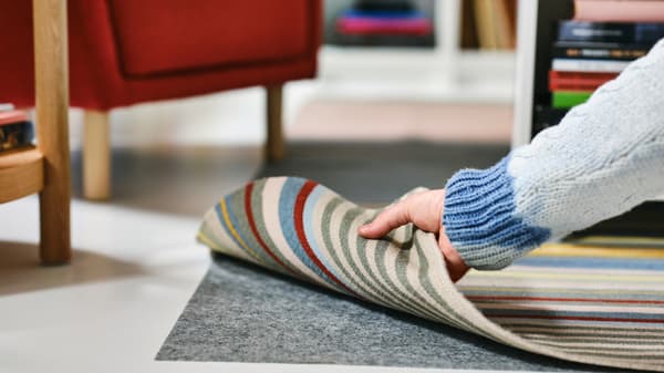 A hand lifts a corner of a multicolour/stripe pattern BUDDINGE flatwoven rug to show STOPP FILT rug underlay with anti-slip.