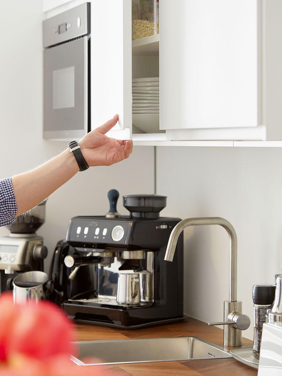 A hand opens a kitchen wall cupboard containing stacked plates. Underneath, one sees a kitchen worktop with sink and mixer tap, next to it a portafilter coffee machine. 