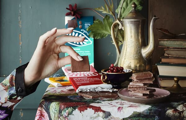 A hand picking up a square of chocolate and a table decorated with chocolate bars, books and fruits.