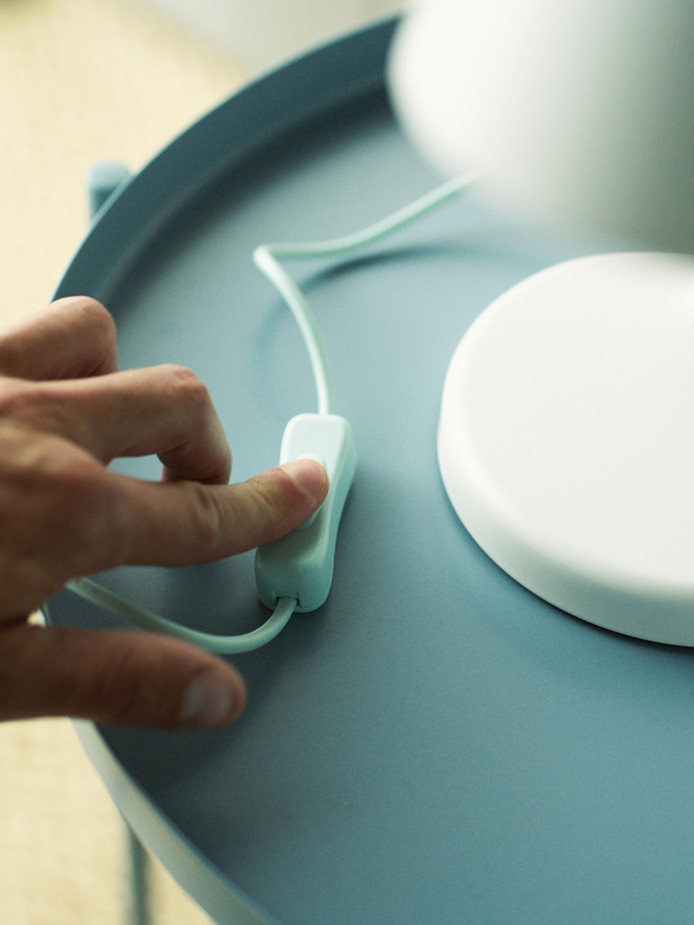 A hand pressing the switch of a white table lamp standing on a light blue table.