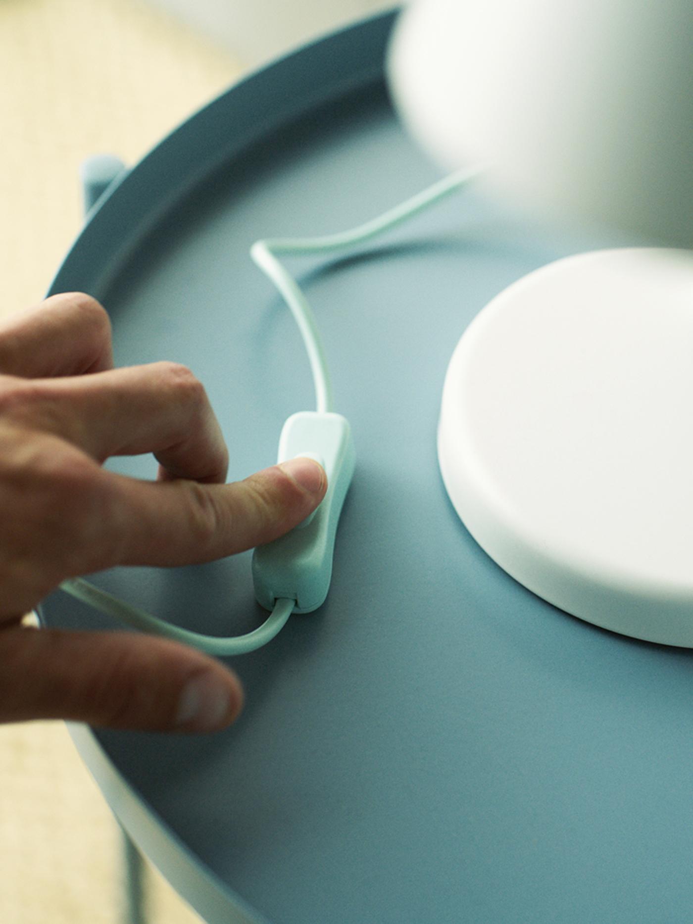 A hand pressing the switch of a white table lamp standing on a light blue table.