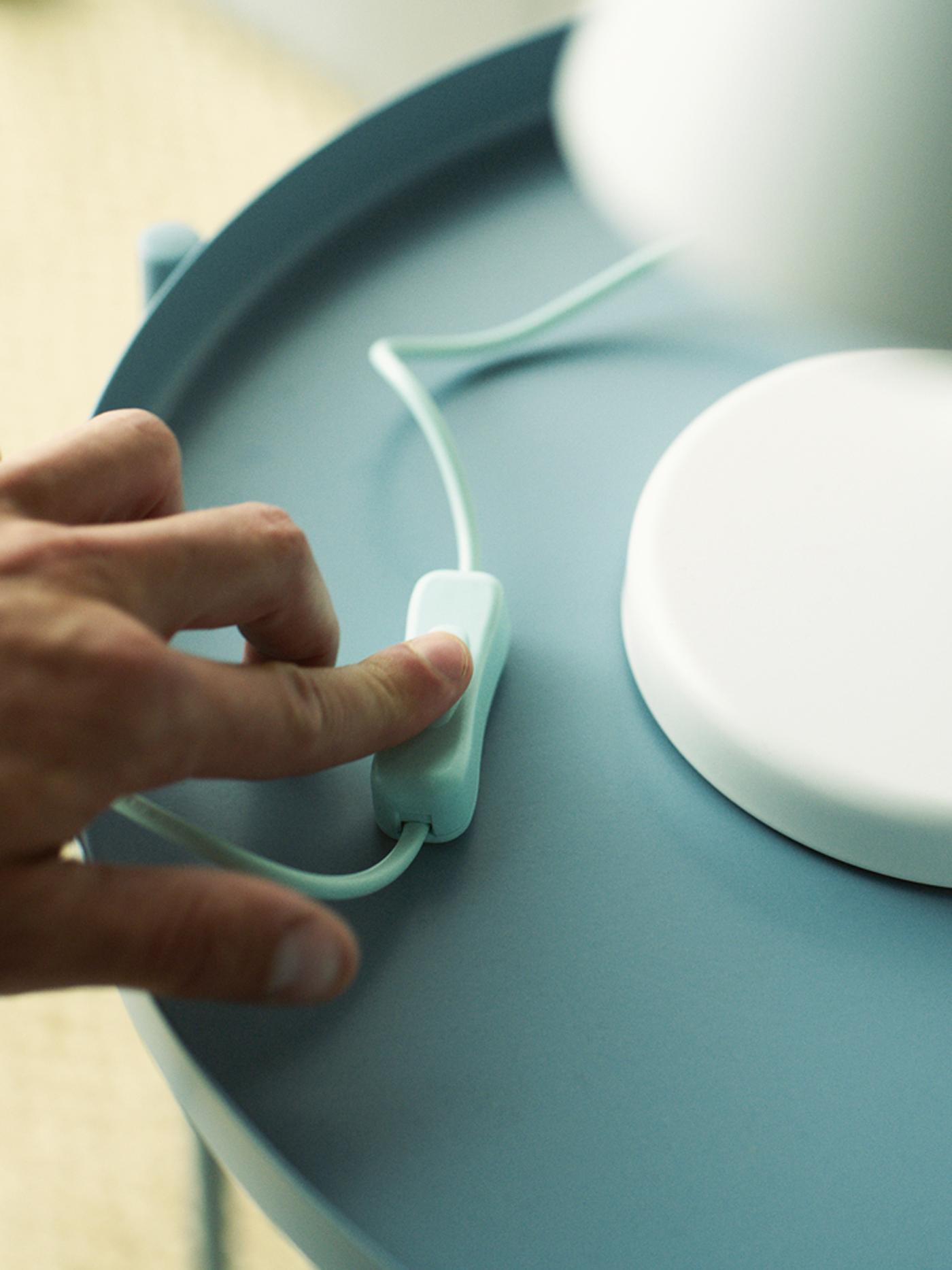 A hand pressing the switch of a white table lamp standing on a light blue table.