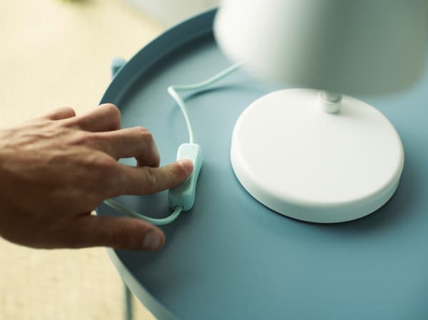 A hand pressing the switch of a white table lamp standing on a light blue table.