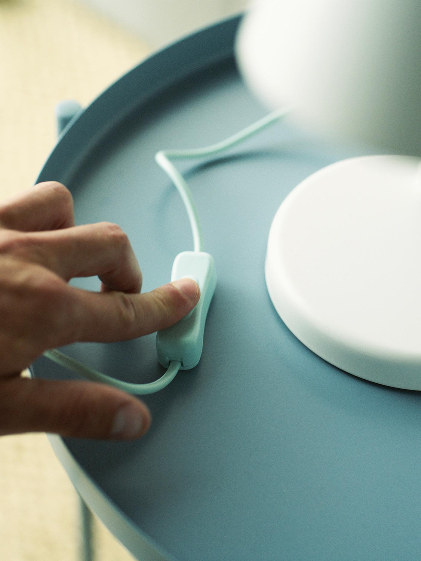 A hand pressing the switch of a white table lamp standing on a light blue table.