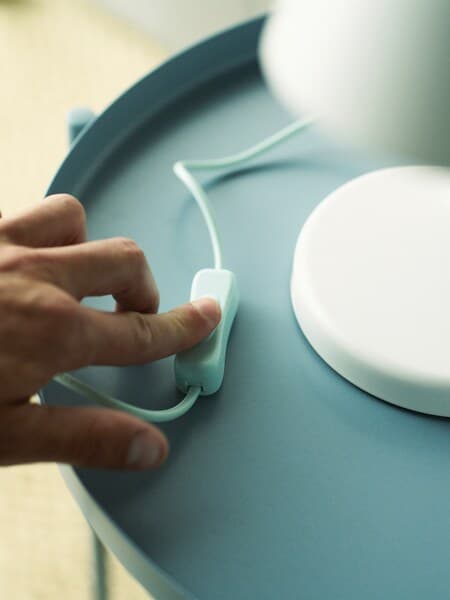 A hand pressing the switch of a white table lamp standing on a light blue table