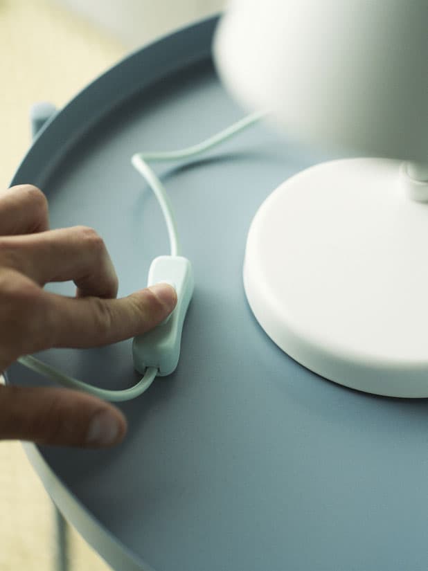 A hand pressing the switch of a white table lamp standing on a light blue table.
