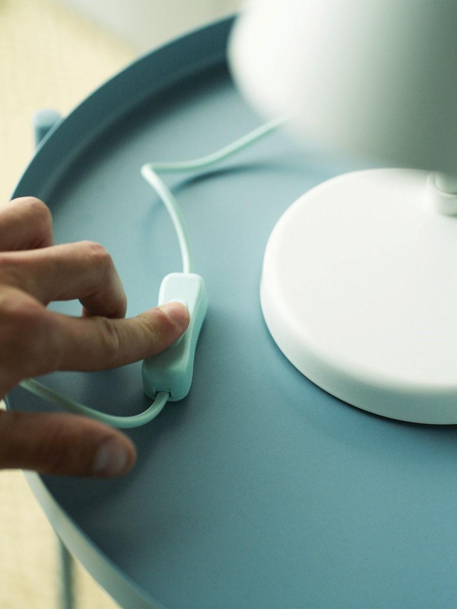 A hand pressing the switch of a white table lamp standing on a light blue table.