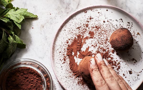 A hand rolling a meatball-sized, round truffle in cocoa powder on a small flat plate; fresh mint leaves on the side.