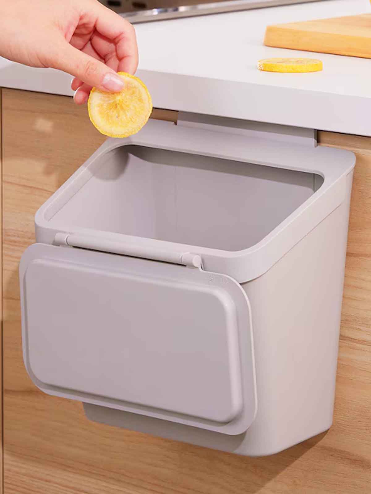 A hand throwing an orange slice into a light grey SKOLÄST waste bin for cabinet with door.